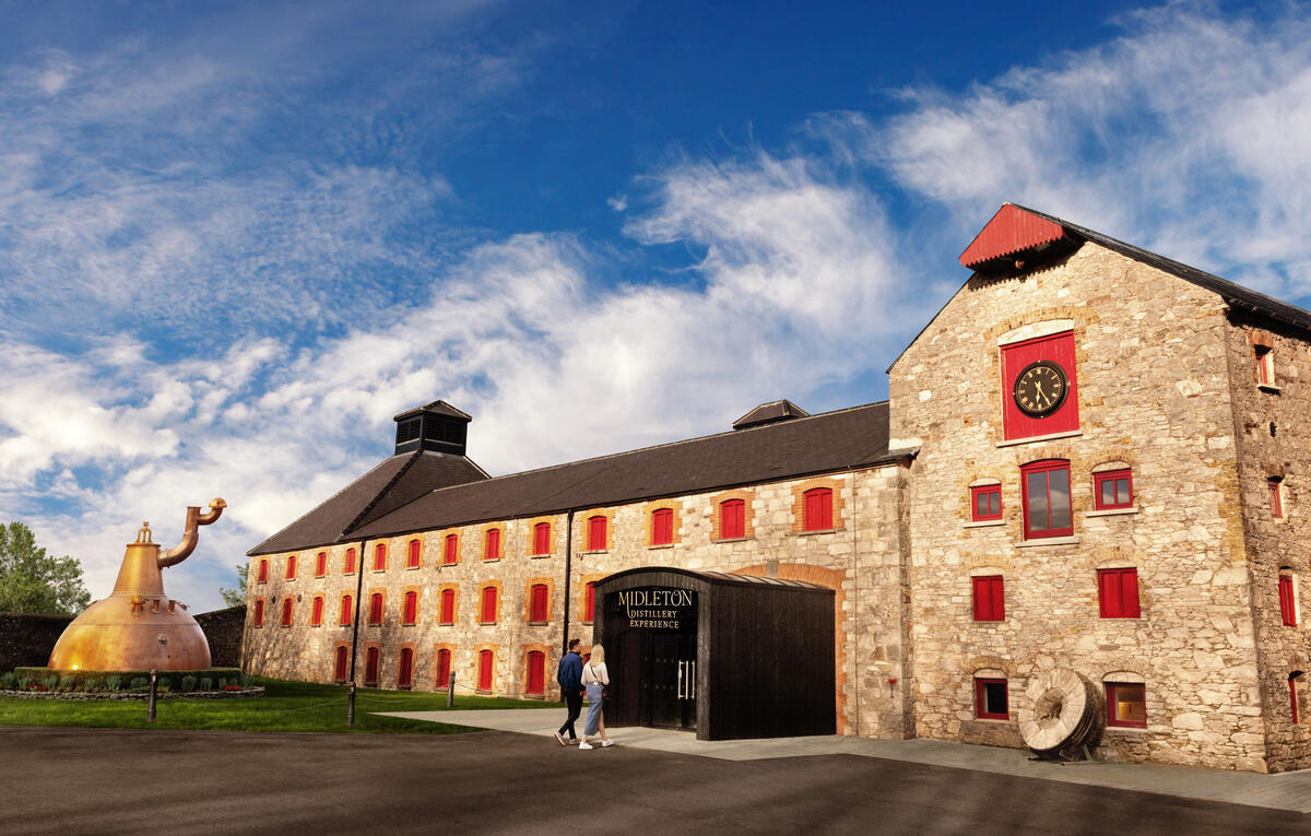 The stone exterior of the Midleton Distillery Experience in County Cork, with red shutters, a large copper pot still sculpture and visitors walking towards the entrance.